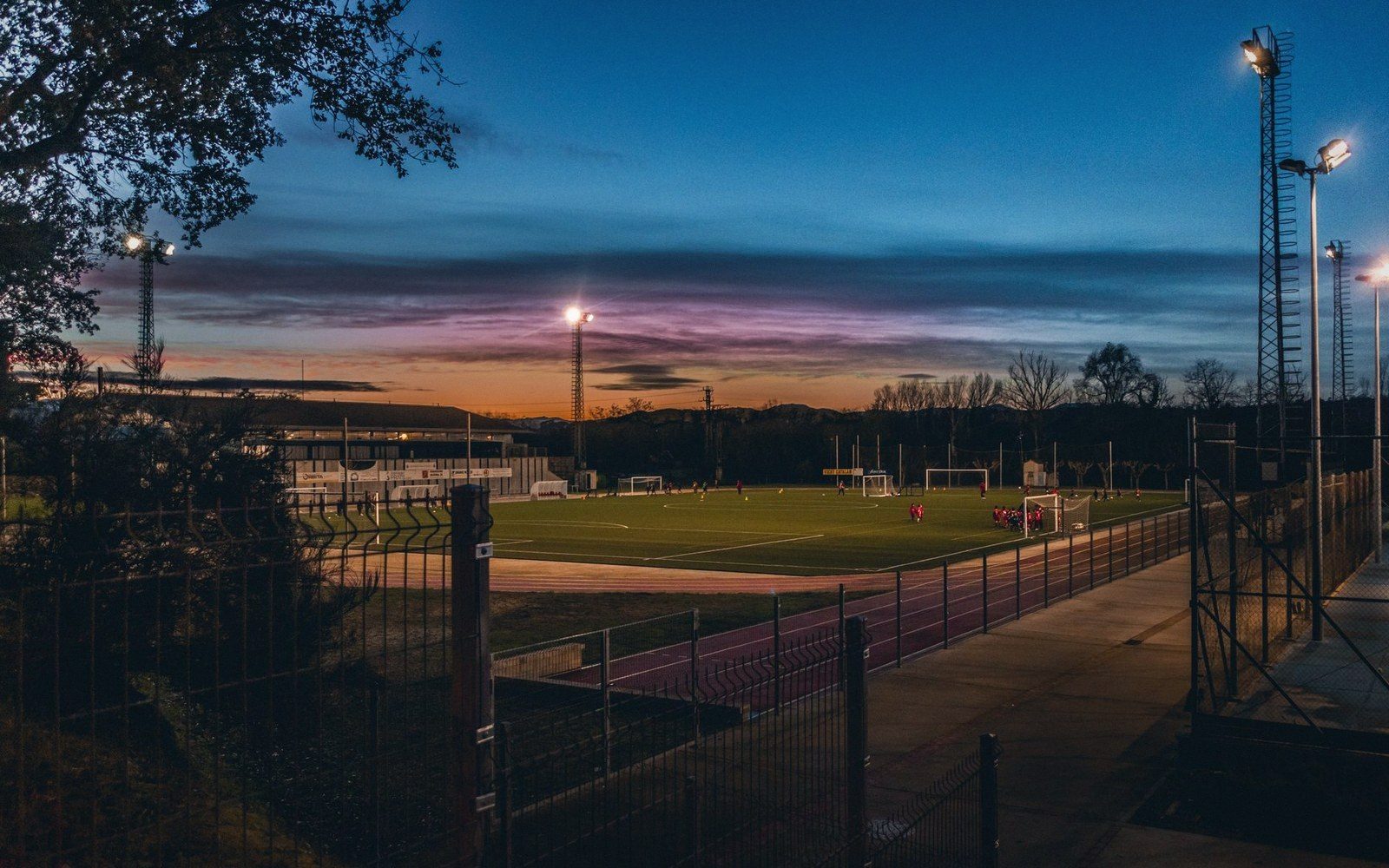 A single groundsman walking the curve of an athletics track, early morning, floodlights still on against grey sky
